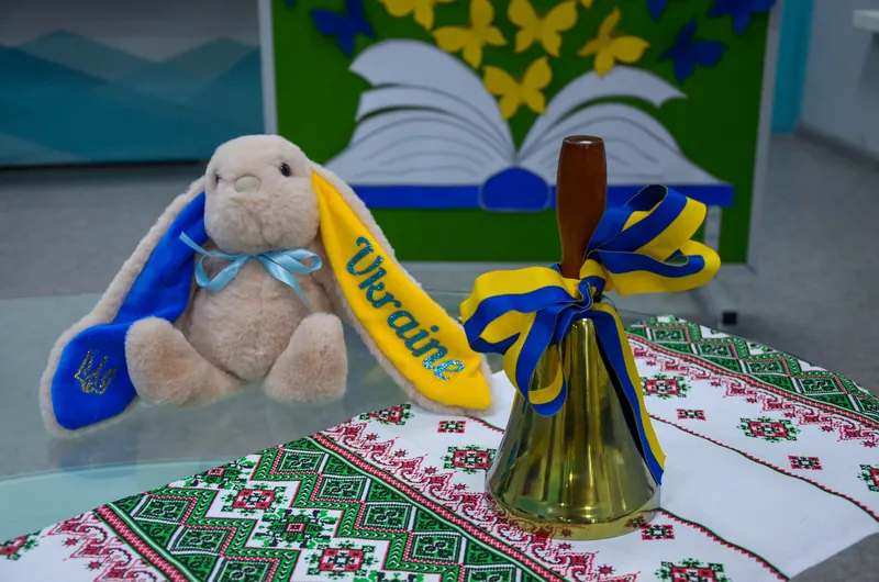 Плюшеве зайченя у синьо-жовтих кольорах і святковий дзвоник зі стрічкою на вишитому рушнику