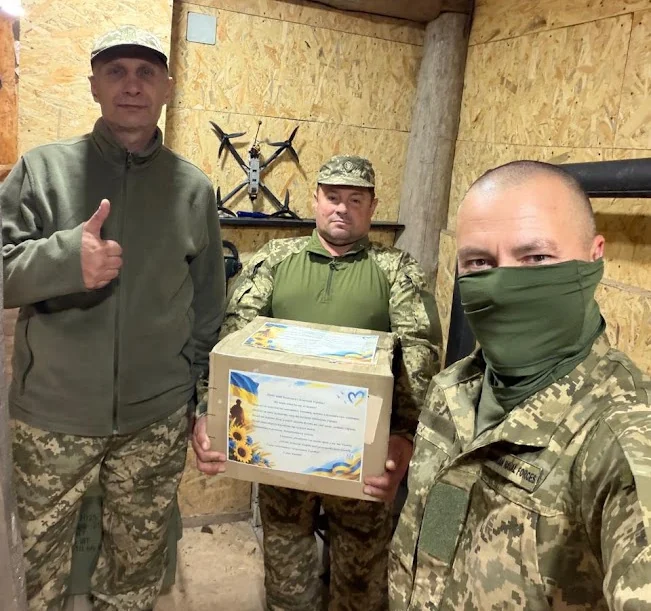 Троє військових у бліндажі тримають картонну коробку з наклеєною листівкою із соняшниками та синьо-жовтим прапором; один показує великий палець угору