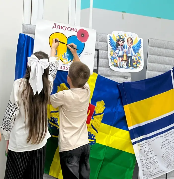 Двоє учнів у вишиванках підписують плакат «Дякуємо за захист» поруч із державними прапорами