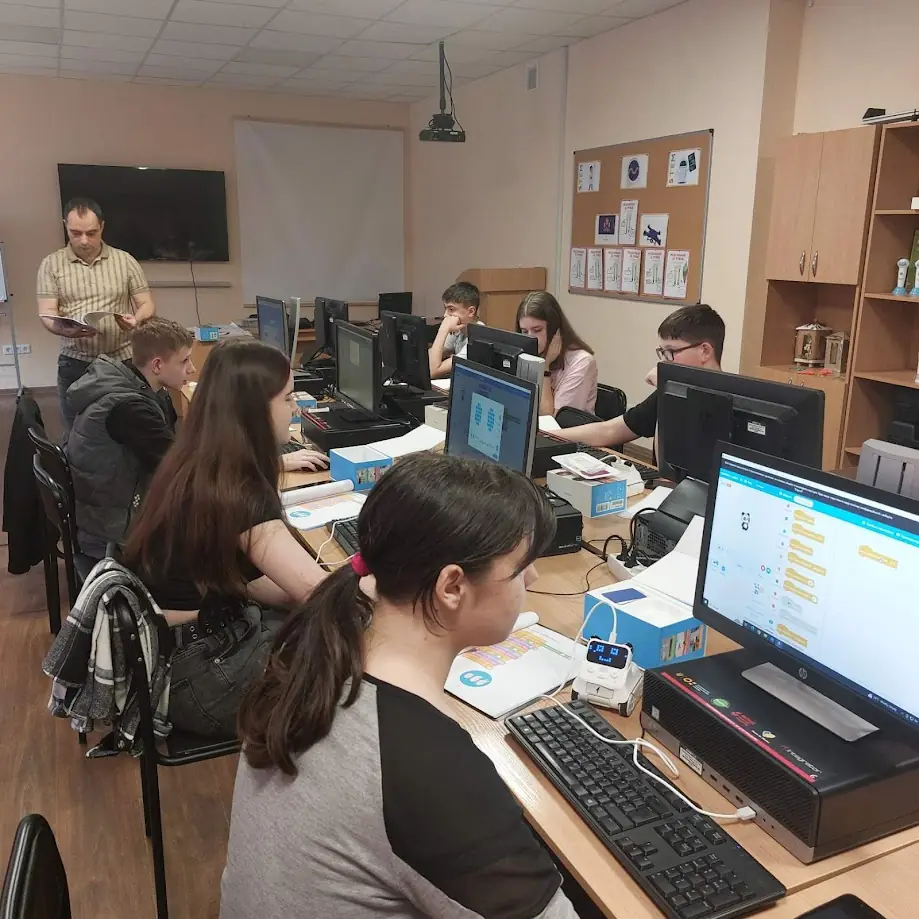 Учні за компʼютерами в кабінеті інформатики програмують навчальних роботів; поруч учитель із журналом